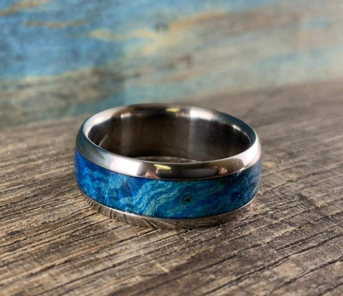 A metal ring with a wide blue wood burl inlay resting diagonally on a rustic wooden surface.