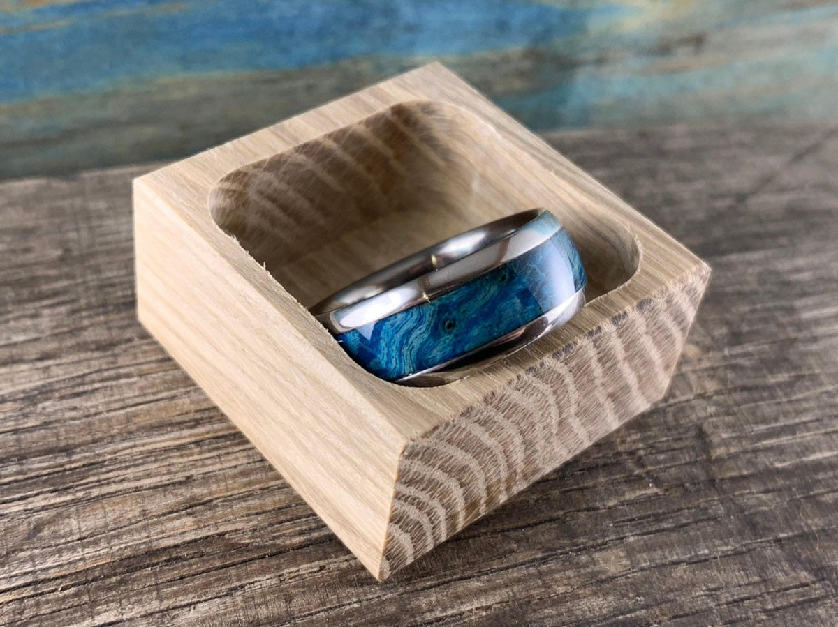 The ring is displayed inside a small, open square wooden box resting on a rustic wooden surface.