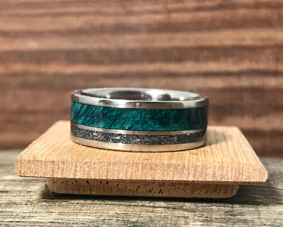 The ring rests on a small square wooden block, displaying the green wood and metallic inlays against a neutral background.