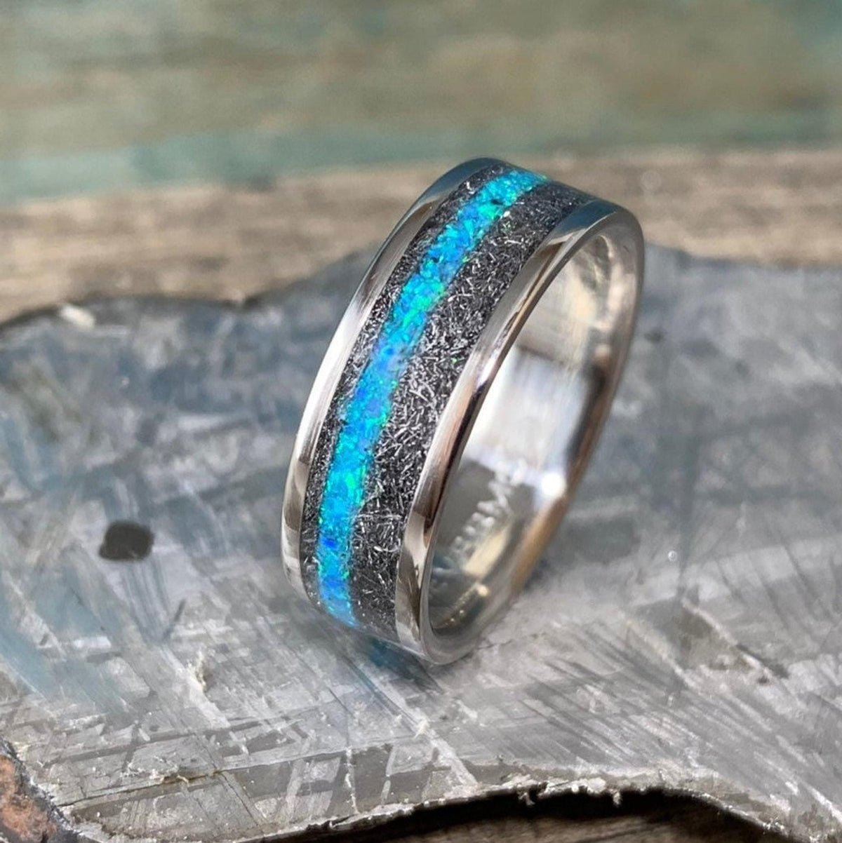 A ring with a meteorite inlay and a blue opal center stripe rests angled on a textured gray slab.