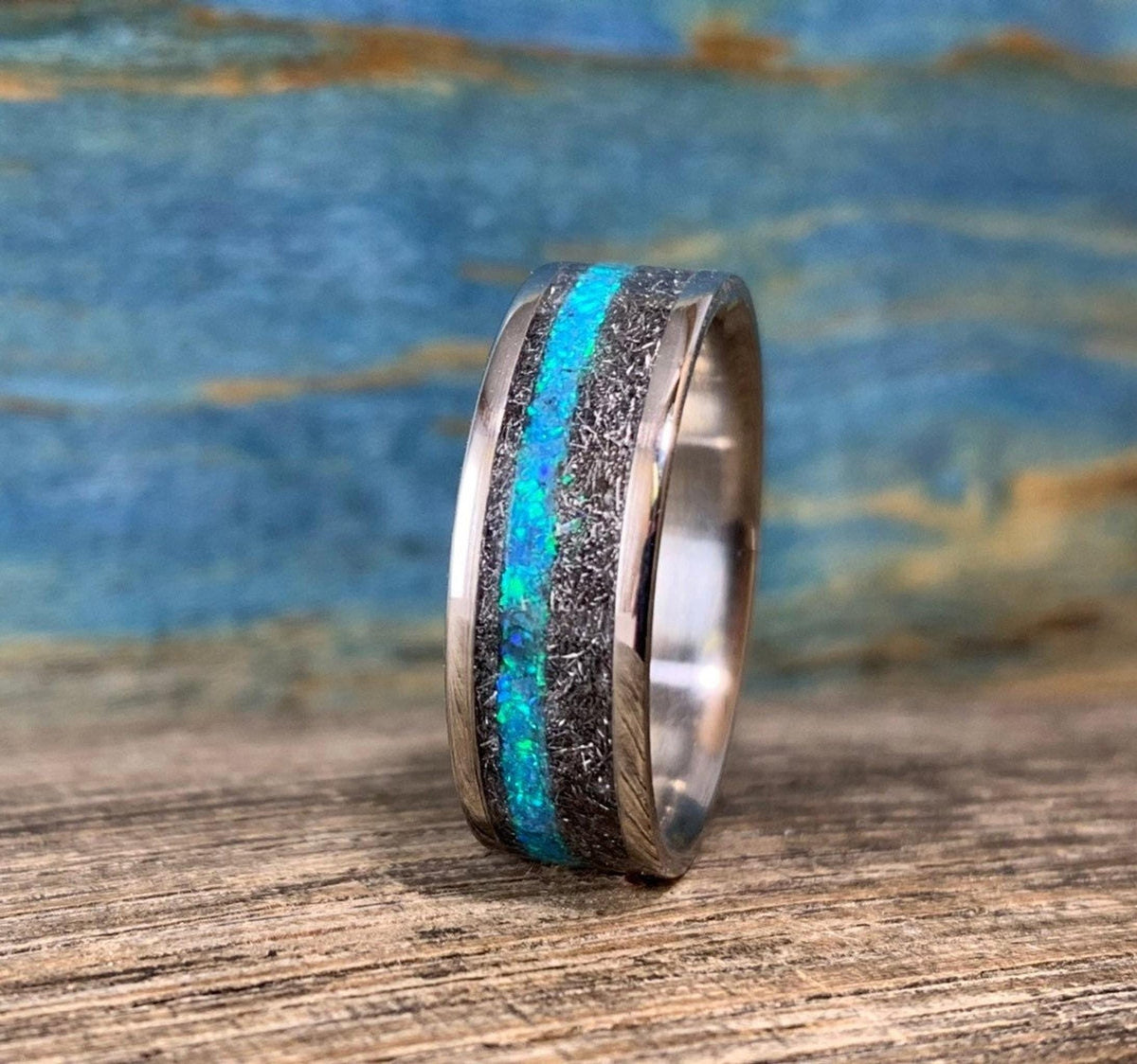 A ring featuring a central blue and green opal inlay set within meteorite and polished metal edges, standing upright on wood.