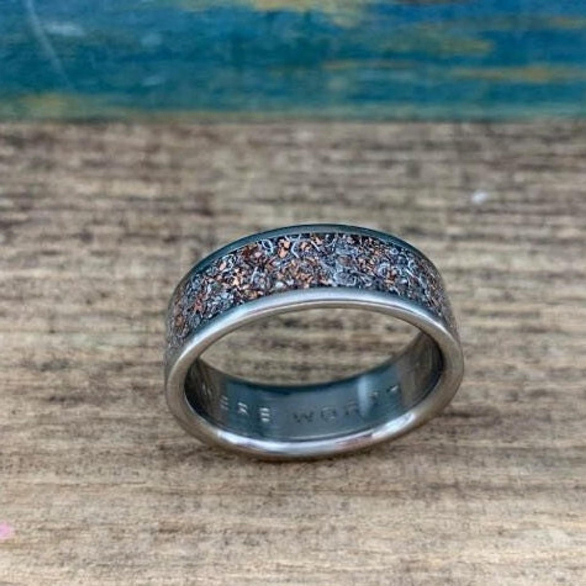 A metal ring lying at an angle, showing its inner band engraving and the textured mixed metal inlay on the outer surface.