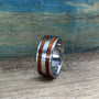 A metal ring with two wood inlays sitting upright on a wood surface against a textured blue and brown background.