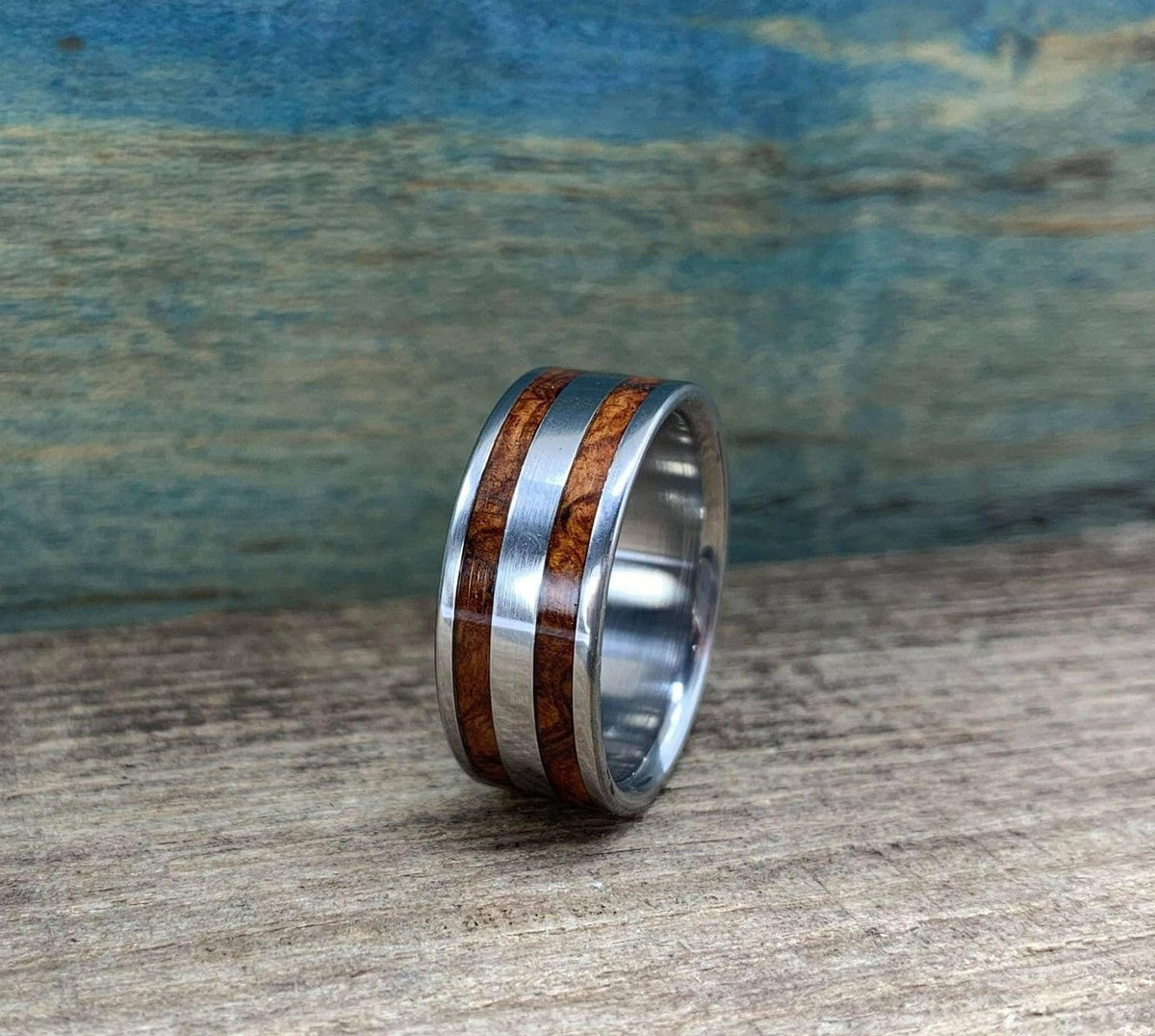 A metal ring with two wood inlays sitting upright on a wood surface against a textured blue and brown background.
