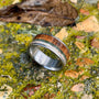 A titanium ring featuring wood, green stone, and antler inlays, resting on mossy rocks with a fallen leaf.