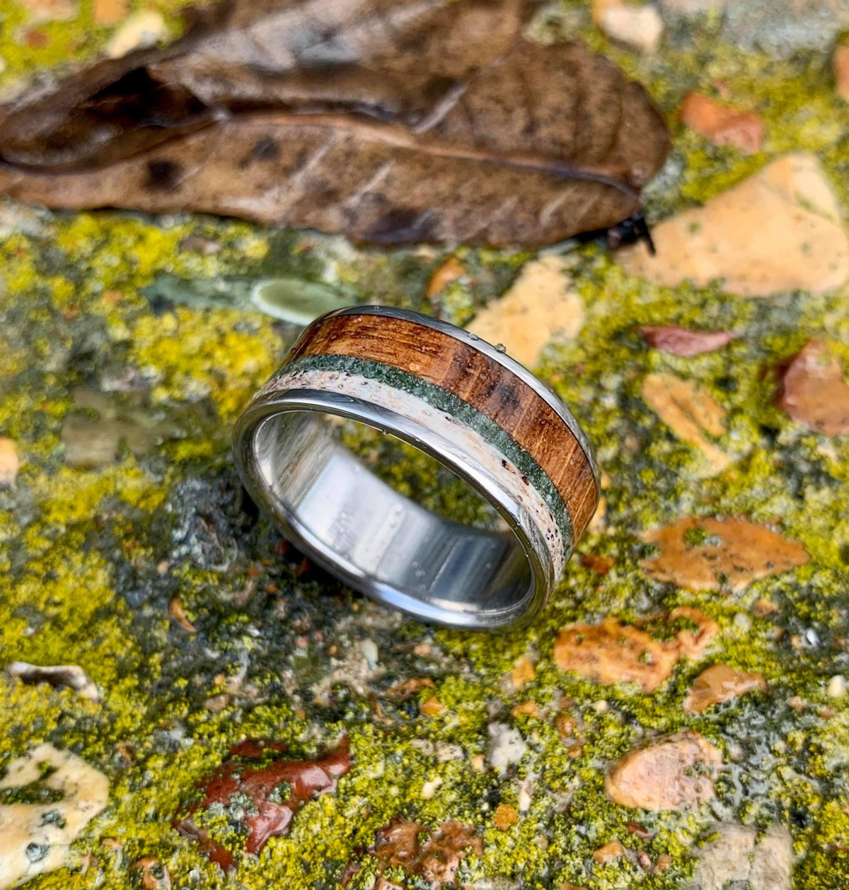A titanium ring featuring wood, green stone, and antler inlays, resting on mossy rocks with a fallen leaf.