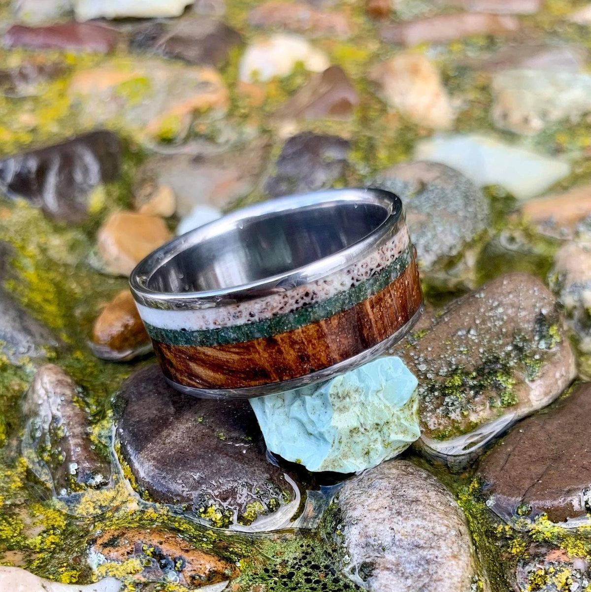 A ring resting on a small turquoise stone, surrounded by wet, moss-covered pebbles.