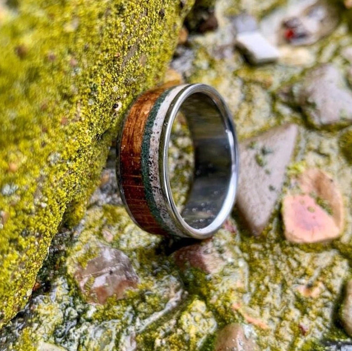 The titanium ring leaning vertically against a mossy green stone surface outdoors.