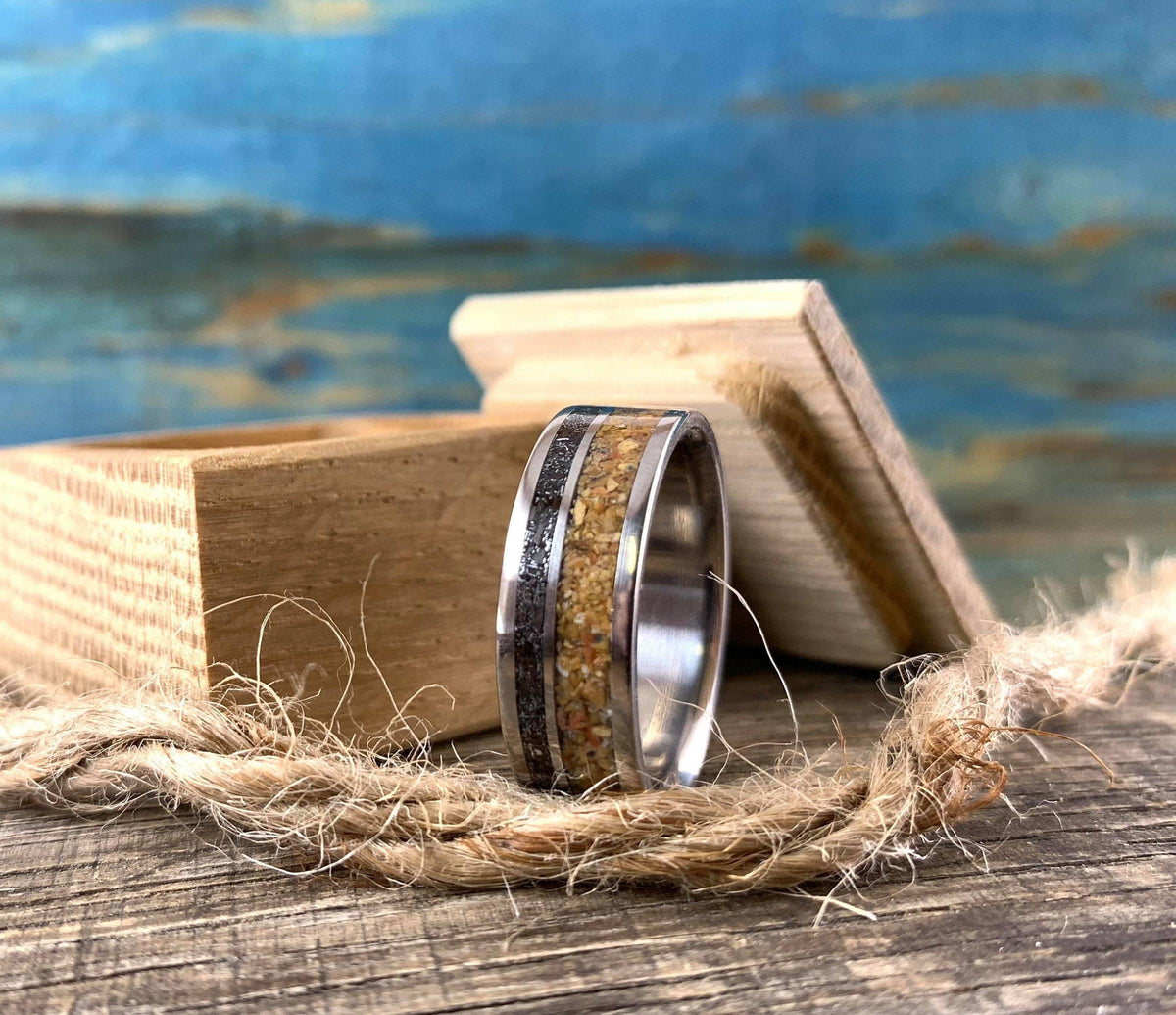 A metal ring with wood and meteorite inlays stands vertically next to an open wooden ring box on a rustic surface.