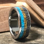 A metal ring with inlays of turquoise, yellow material, and dark meteorite rests on a rustic wooden surface next to an open wooden box.