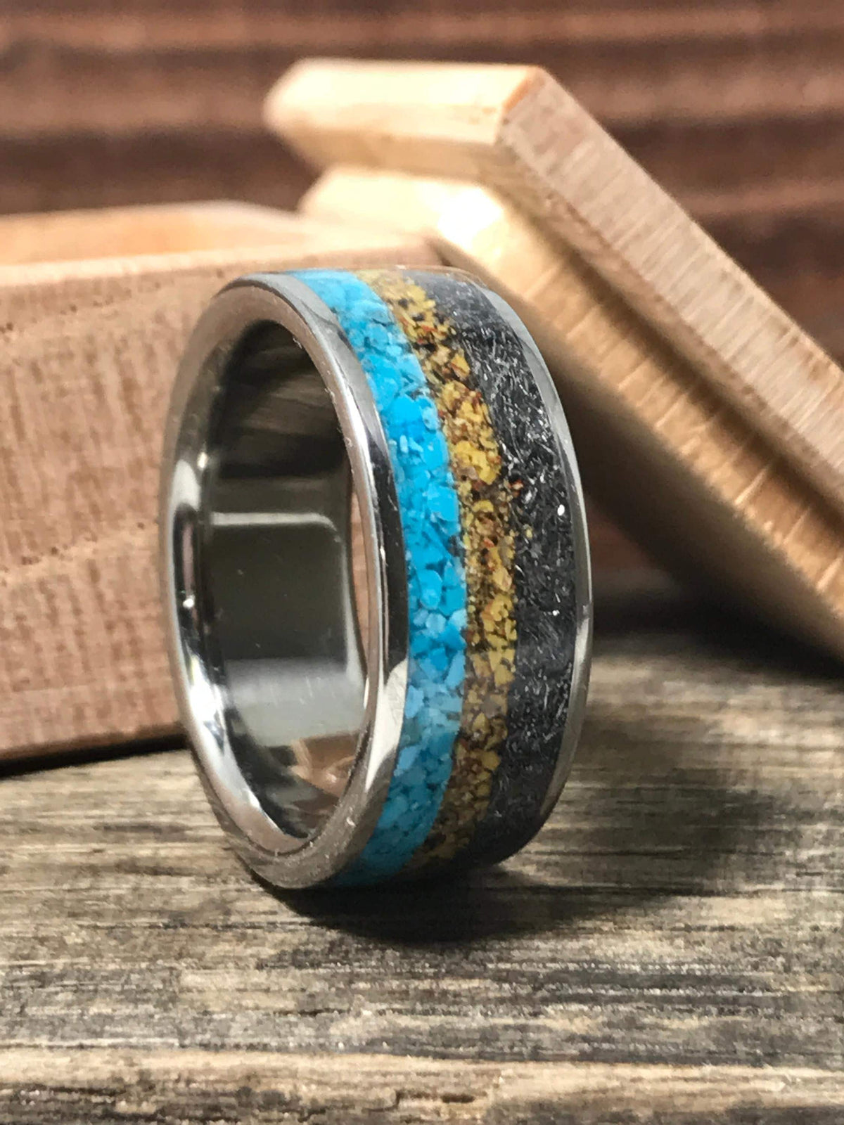 A metal ring with inlays of turquoise, yellow material, and dark meteorite rests on a rustic wooden surface next to an open wooden box.