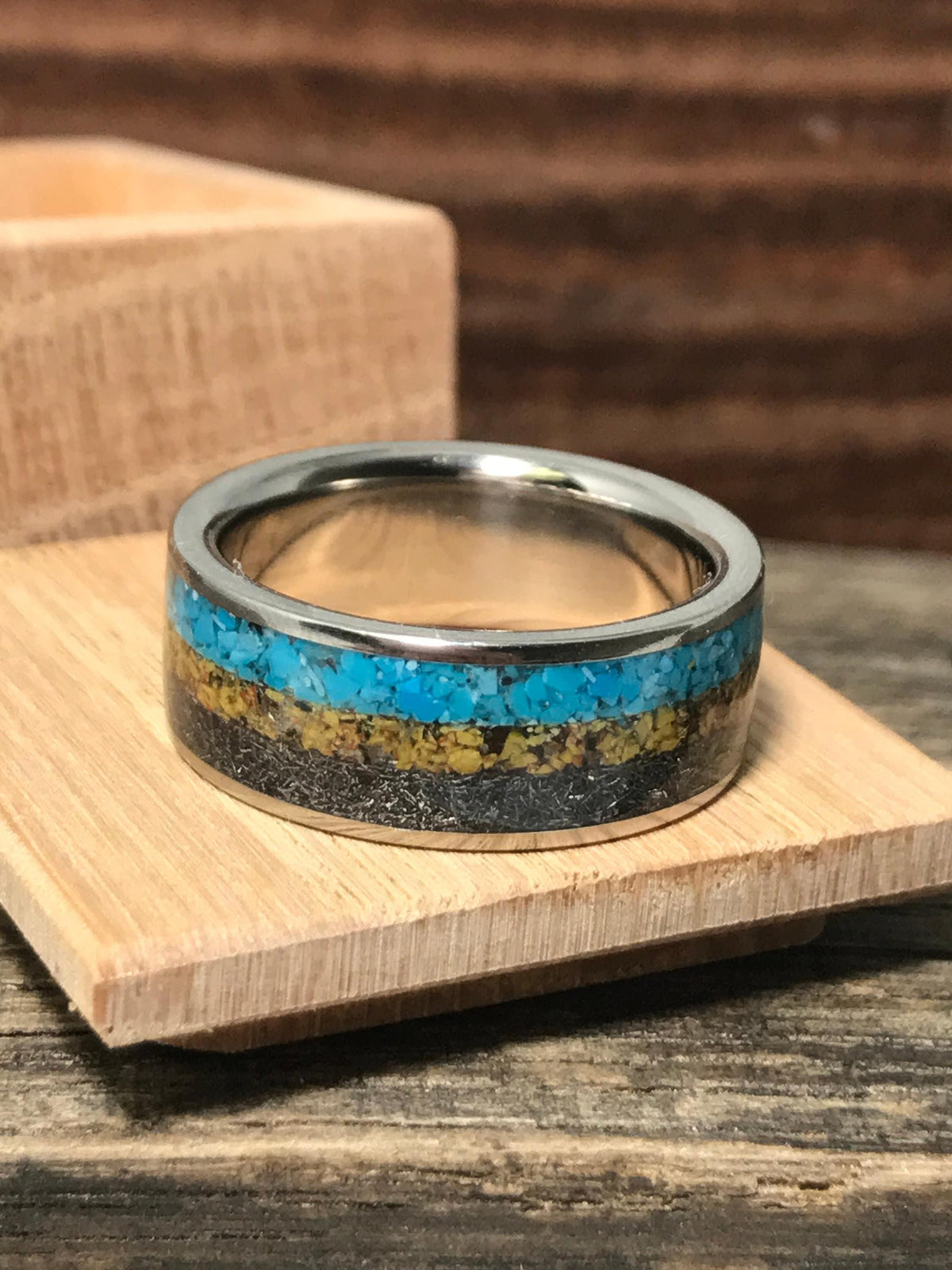 A metal wedding band lies flat on a small square wooden platform, showing horizontal bands of turquoise, yellow, and dark fragments.