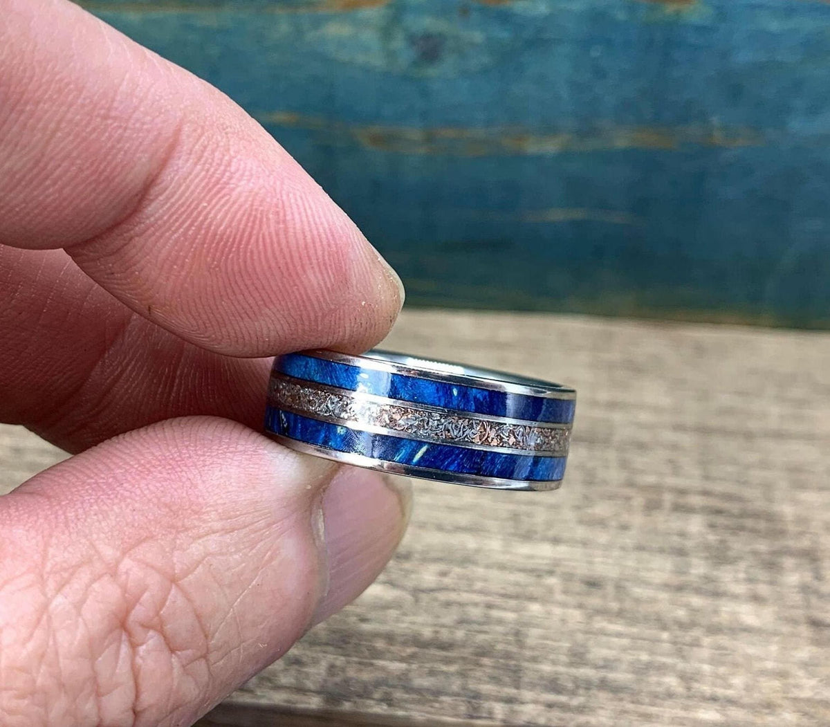 A close-up view of fingers holding the ring horizontally, showcasing its blue and metallic patterned band.