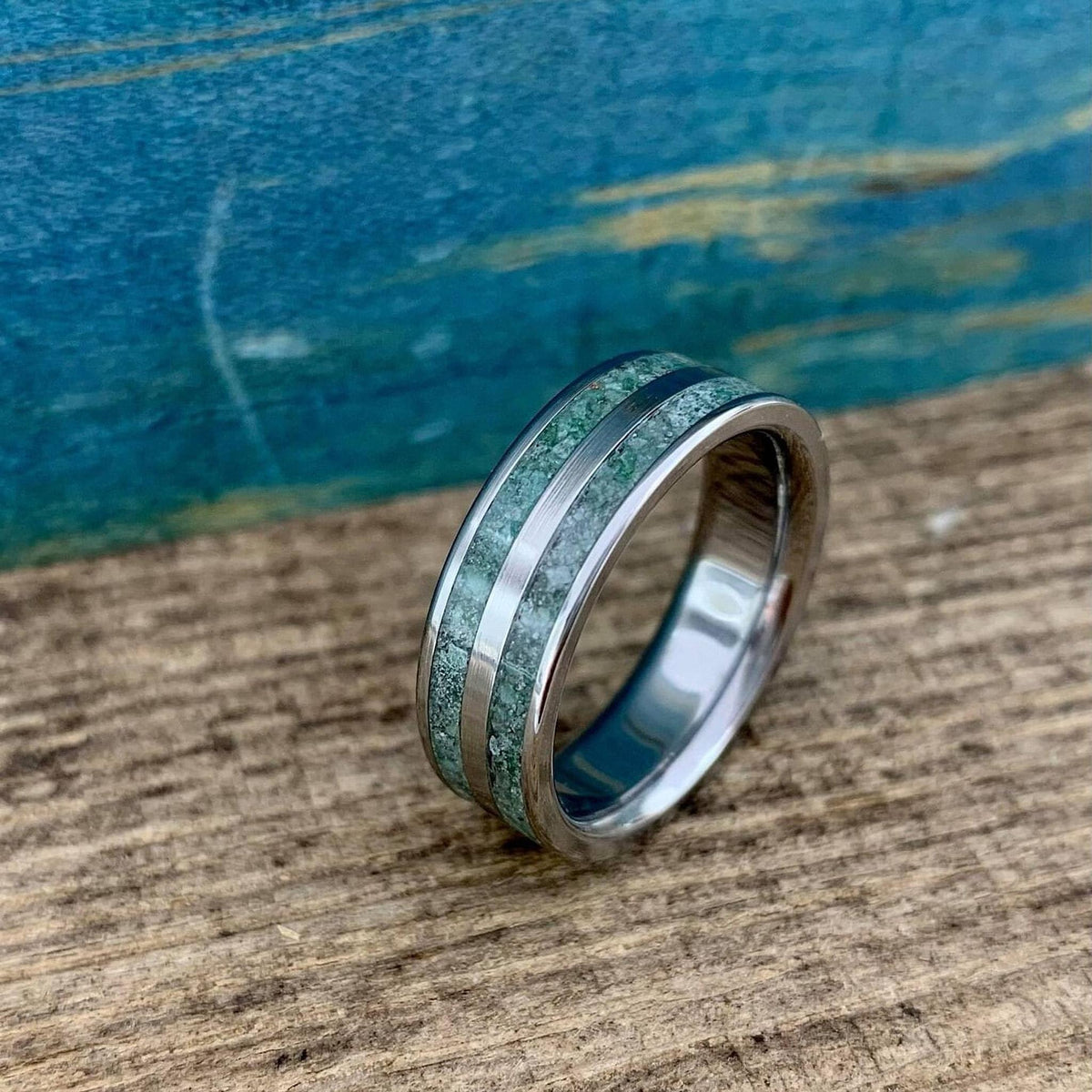 A titanium ring with two green moss agate inlay tracks is positioned at an angle on a textured wooden board.