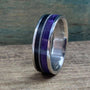A titanium ring featuring an inlay of black ebony, a metal guitar string, and purple box elder wood, shown at an angle on a wooden surface.