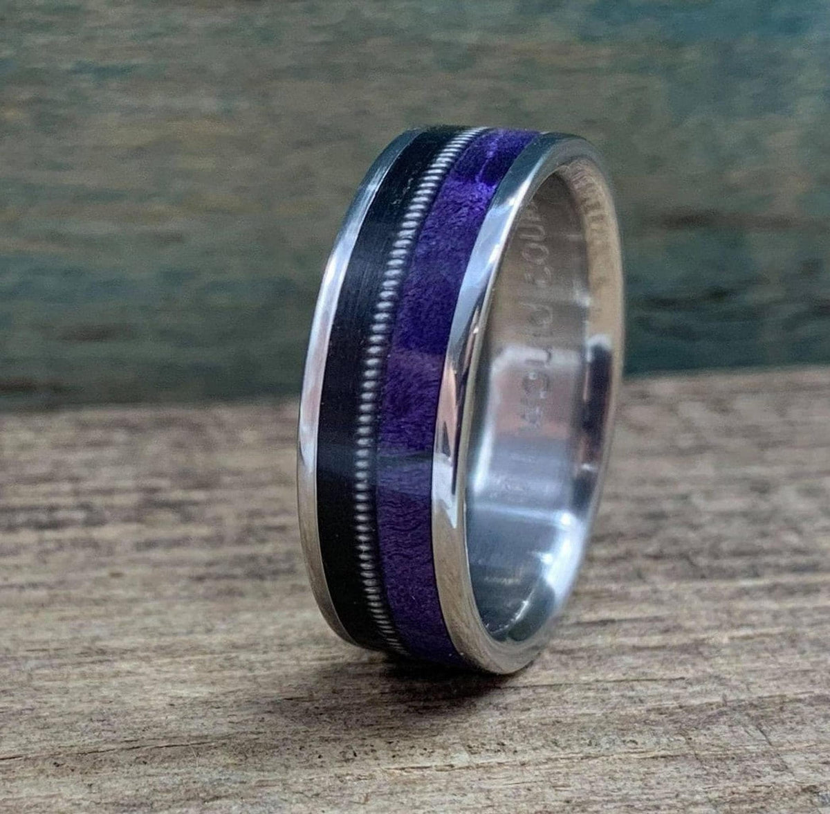 A titanium ring featuring an inlay of black ebony, a metal guitar string, and purple box elder wood, shown at an angle on a wooden surface.