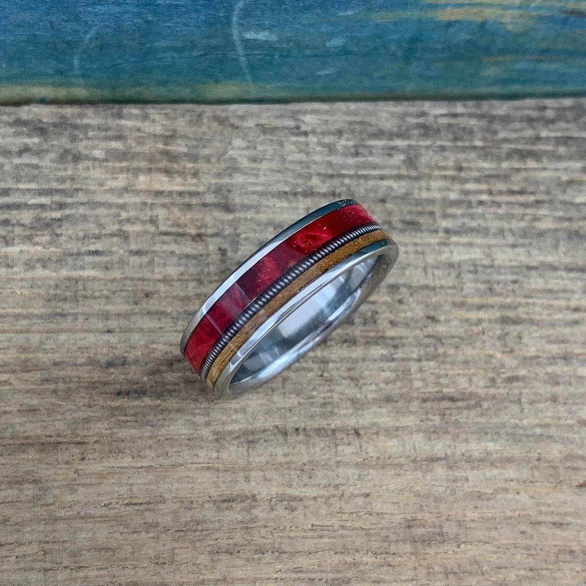 View of the ring lying at a slight angle on a textured wood background, displaying the segmented red wood blocks alongside the metal string and brown wood.