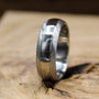 A silver metal ring with a brushed texture and a polished center groove resting upright on a rustic wooden block.