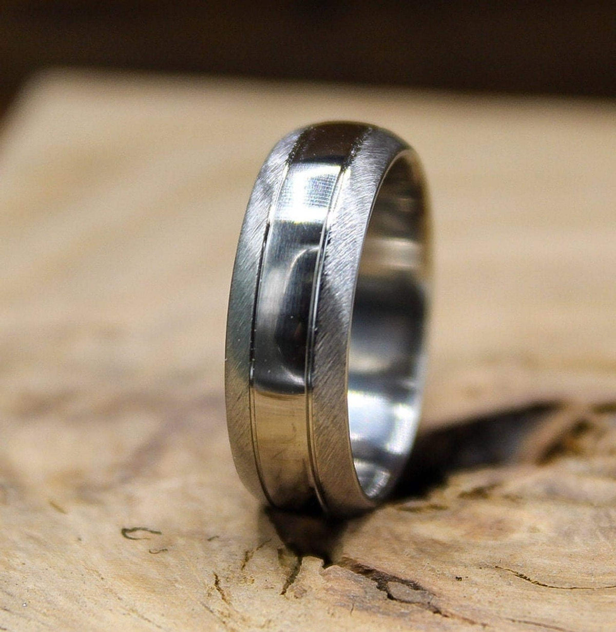 A silver metal ring with a brushed texture and a polished center groove resting upright on a rustic wooden block.