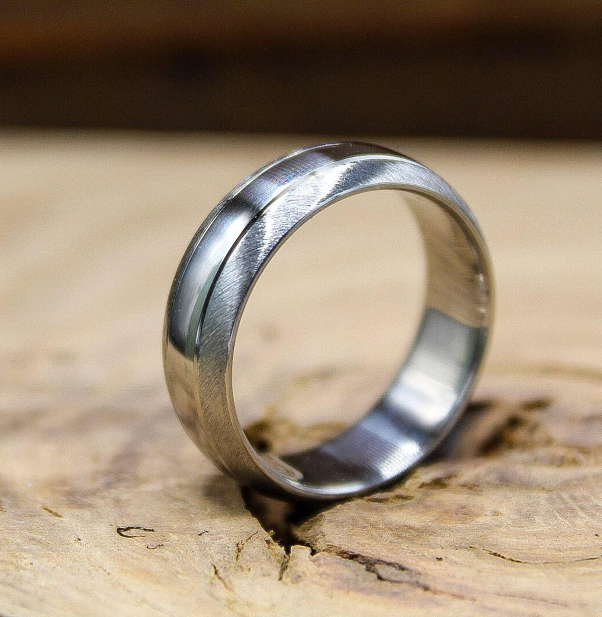 A silver ring with a brushed matte finish and a thin polished groove running around it, angled on a piece of wood.