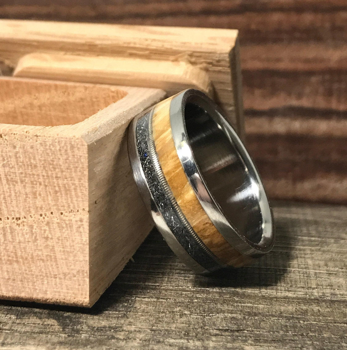 A band ring with metal, wood, and meteorite inlays leaning against an open wooden box on a rustic wood surface.
