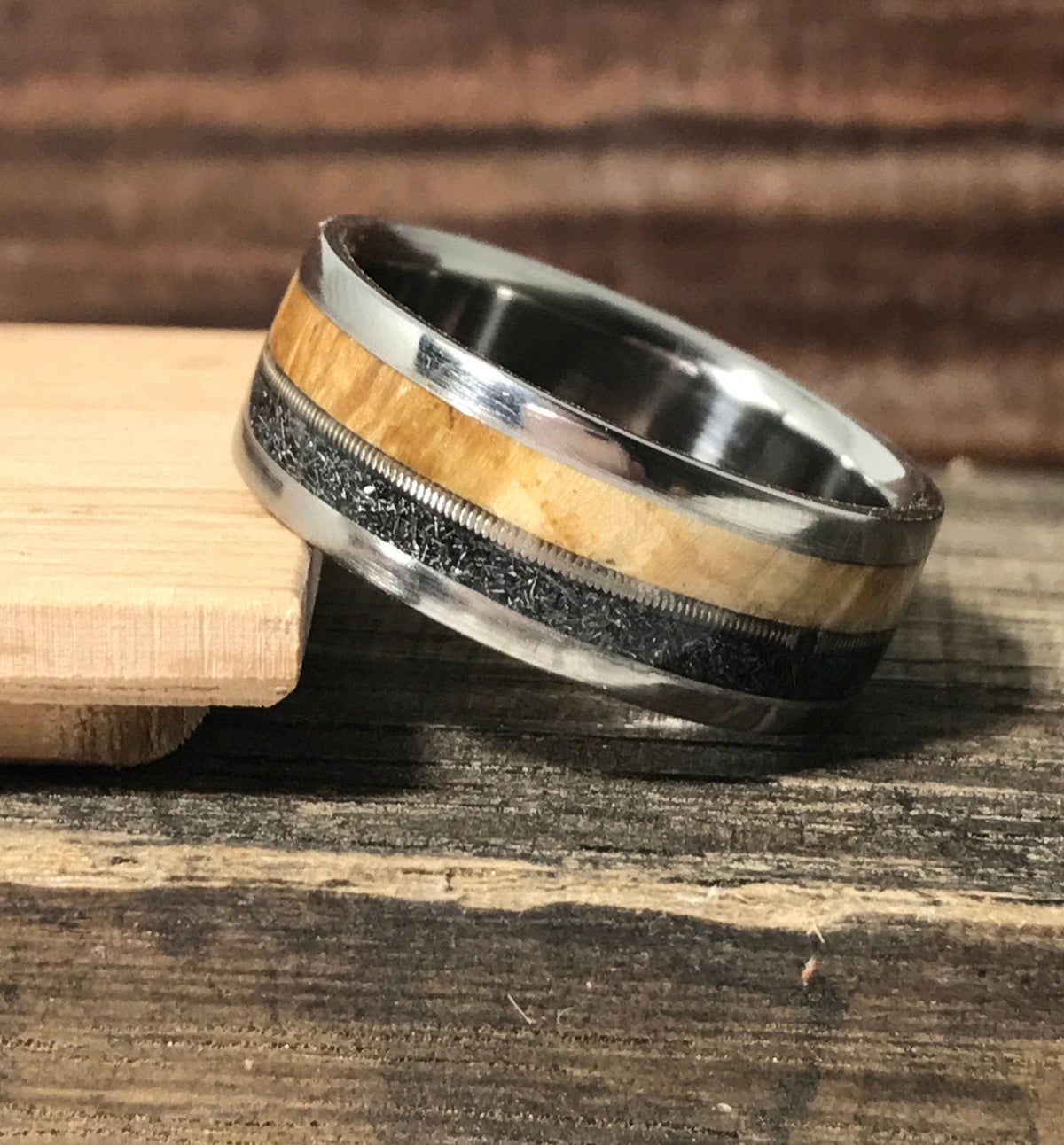 A multi-material ring resting on a small wooden tray against a dark wood background.