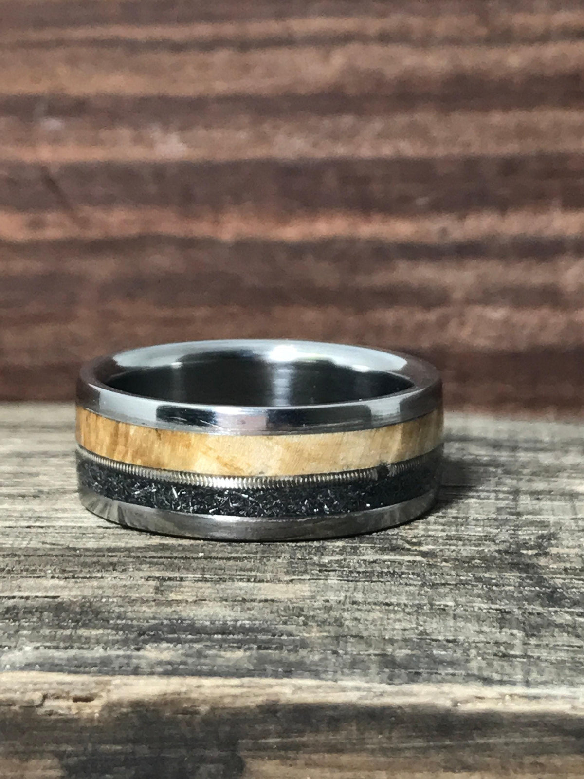 A titanium ring with wooden and dark textured inlays lying horizontally on a weathered wooden plank.