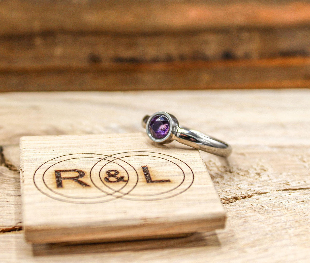 A silver-colored ring with a round purple gemstone in a bezel setting resting next to a small wooden block engraved with letters R and L.