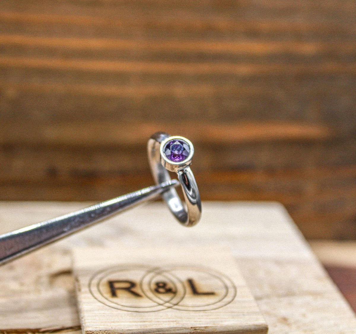 A pair of metal tweezers holding a silver-colored ring featuring a bezel-set purple gemstone, with an engraved wooden block in the blurred background.