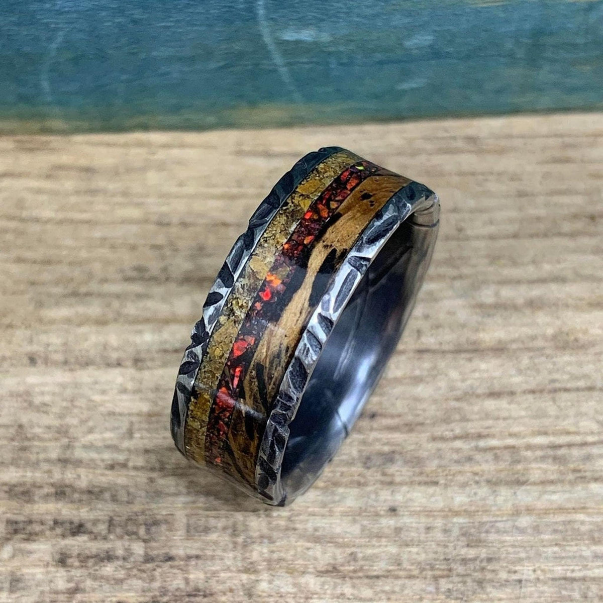 A textured metal wedding band with wood, stone, and opal inlay rests angled on a light wood surface against a teal background.