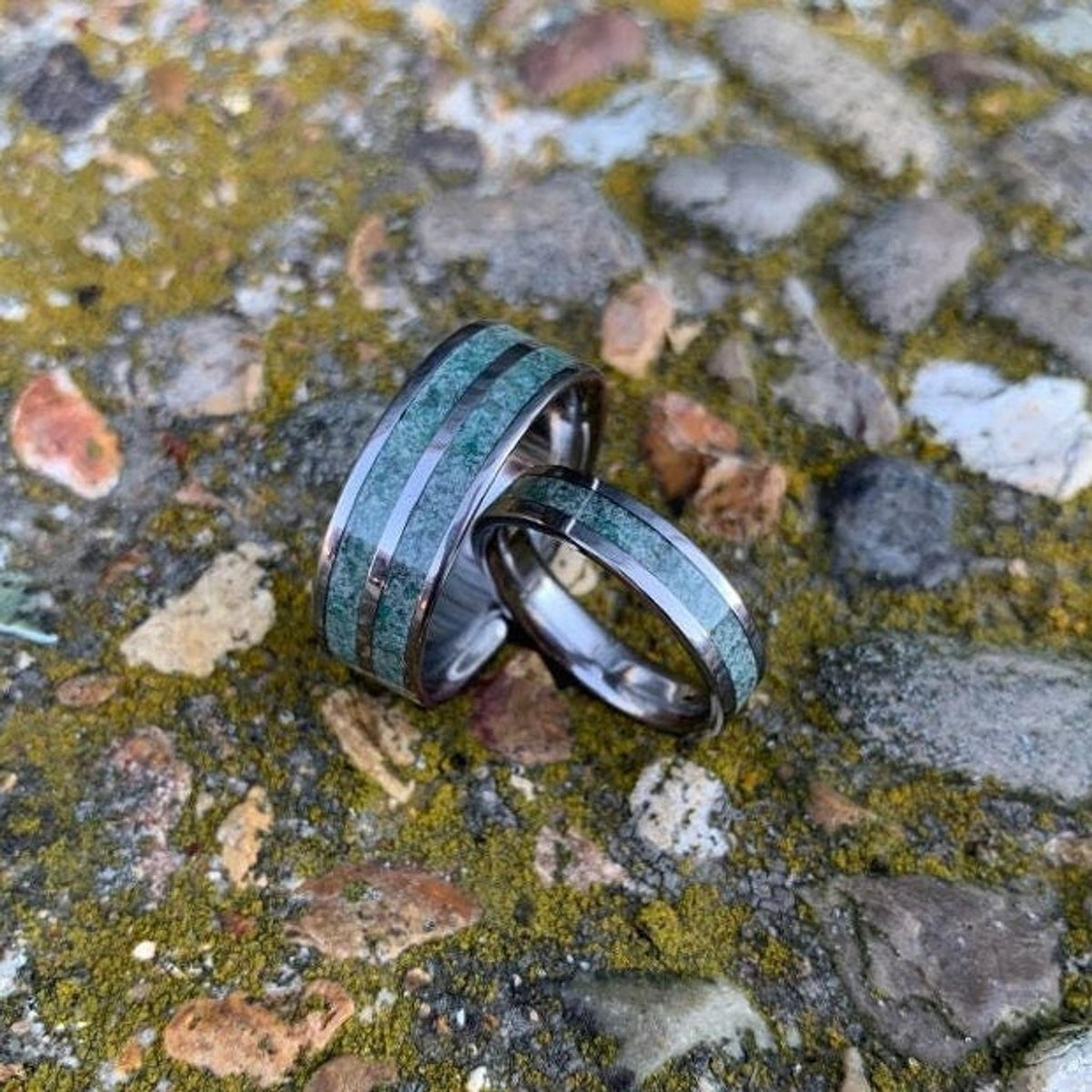 Two titanium rings featuring inlaid green moss agate sit closely together on a mossy, rocky background.