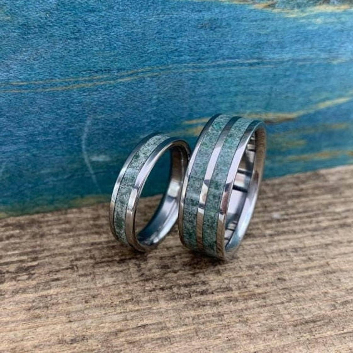 Two titanium bands with green stone inlays sit on a rustic stone and moss background.