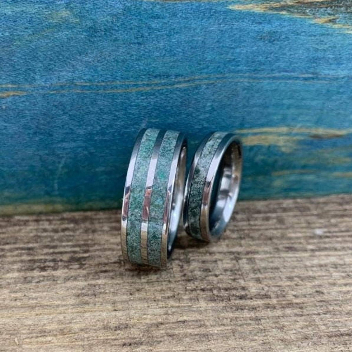Two silver-toned rings with green moss agate inlays stand on a textured wooden surface against a blue-toned backdrop.