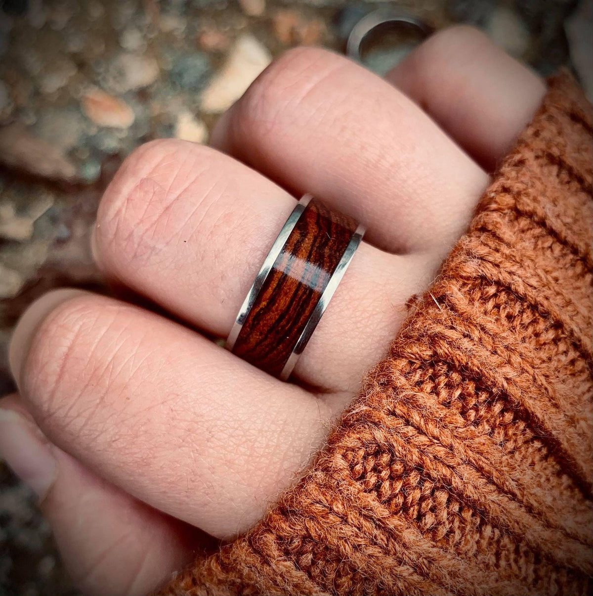 A hand wearing a ring with a wooden inlay and a rust colored sweater.