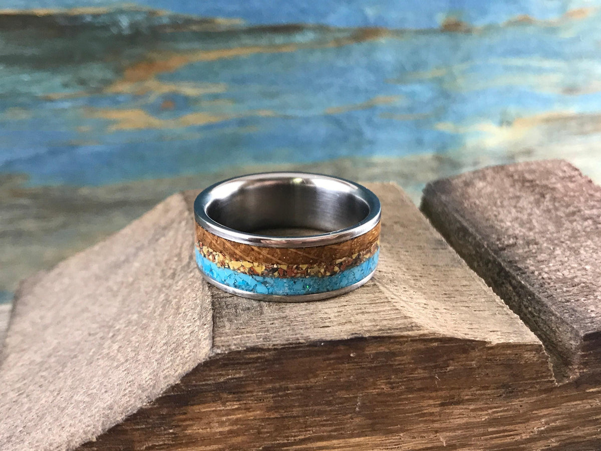 A handmade ring with multicolored inlays sits on a rustic wooden platform with a painted blue backdrop.