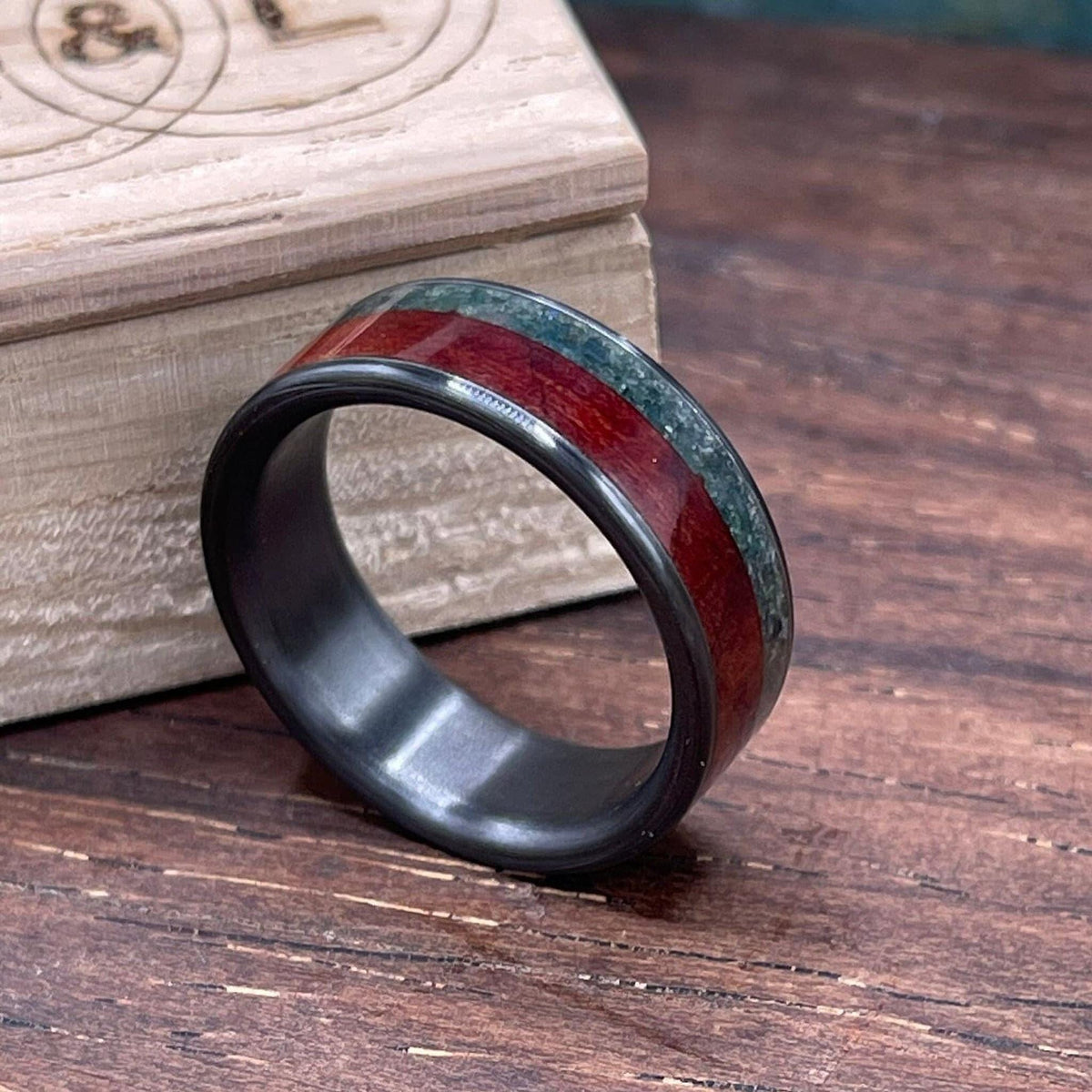 A black band ring with an inlay of red wood and green moss agate rests on a wooden surface next to an engraved wooden box.