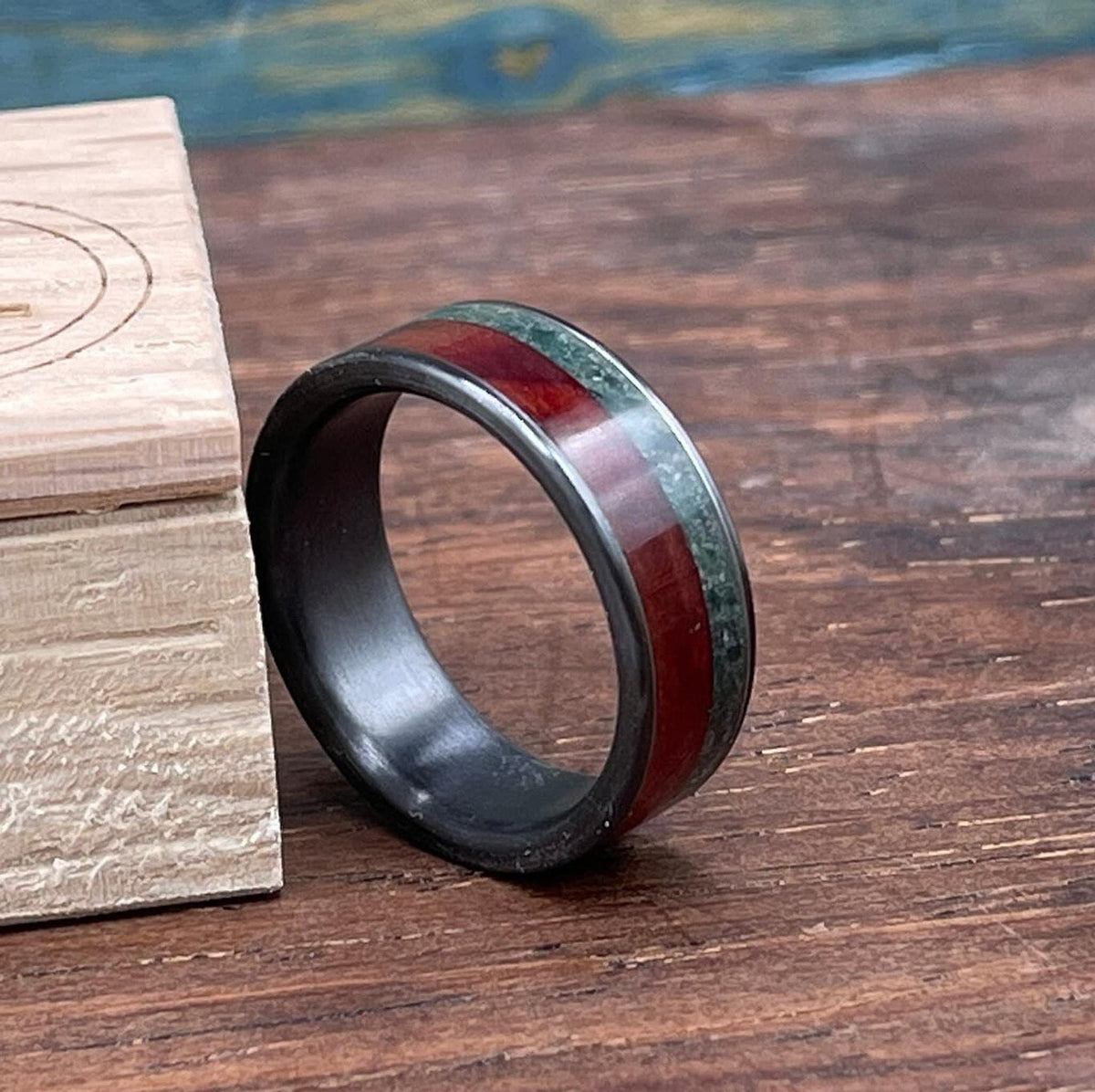 A close-up of the black titanium ring showing its red wood and green moss agate inlay sections next to a wooden box lid.