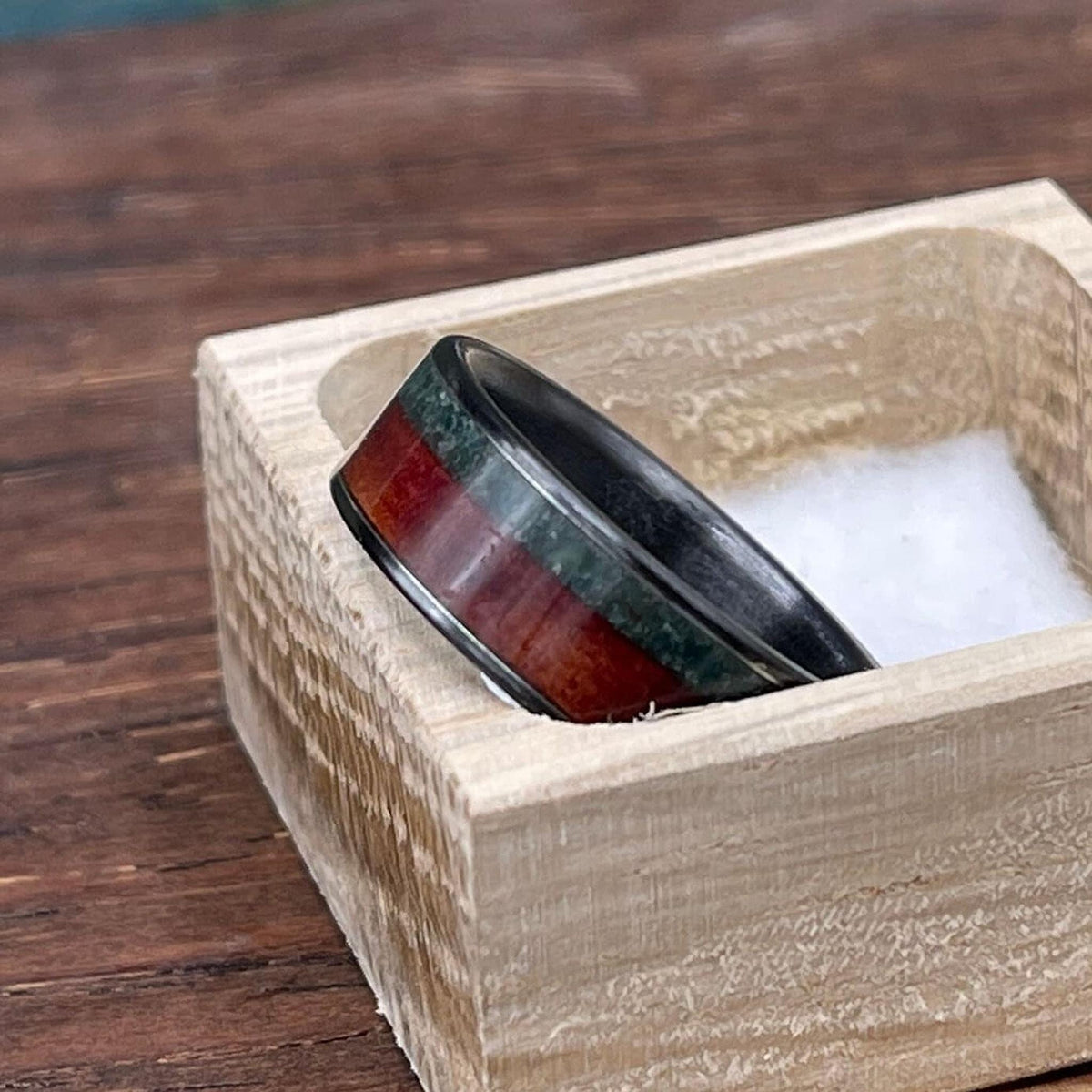 The ring sits inside an open square wooden box lined with white material on a dark wooden table.