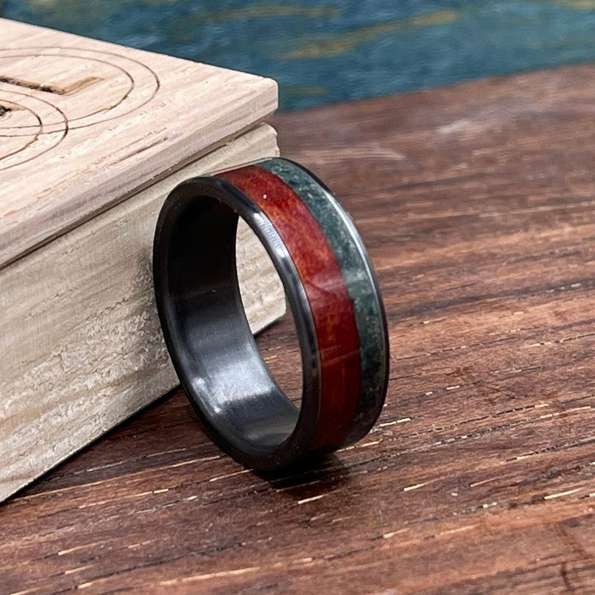 A side view of the black band with a red wood and green moss agate inlay standing on a wooden table near a small box.