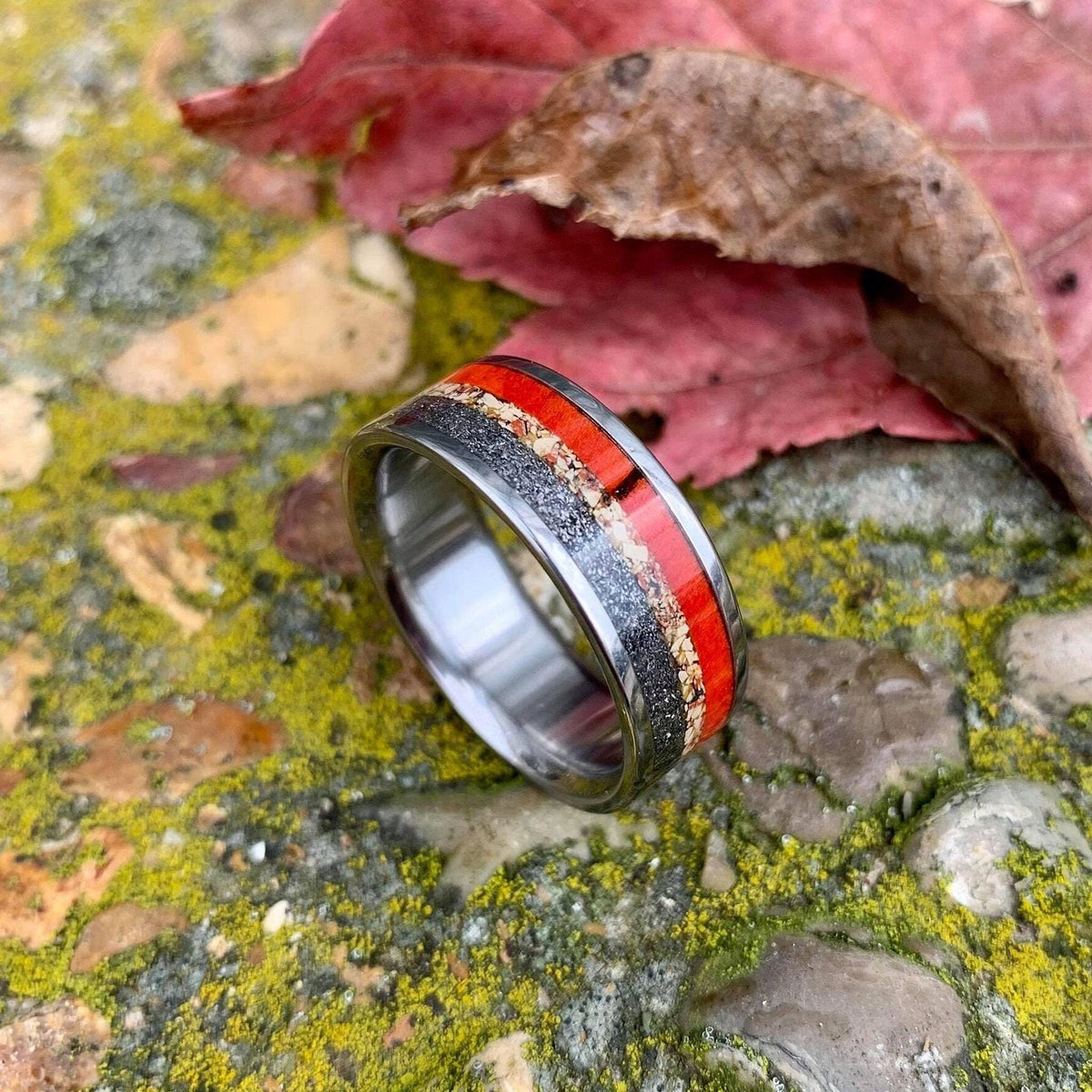 The multi-material inlay ring positioned upright in front of a fallen red leaf on a mossy surface.