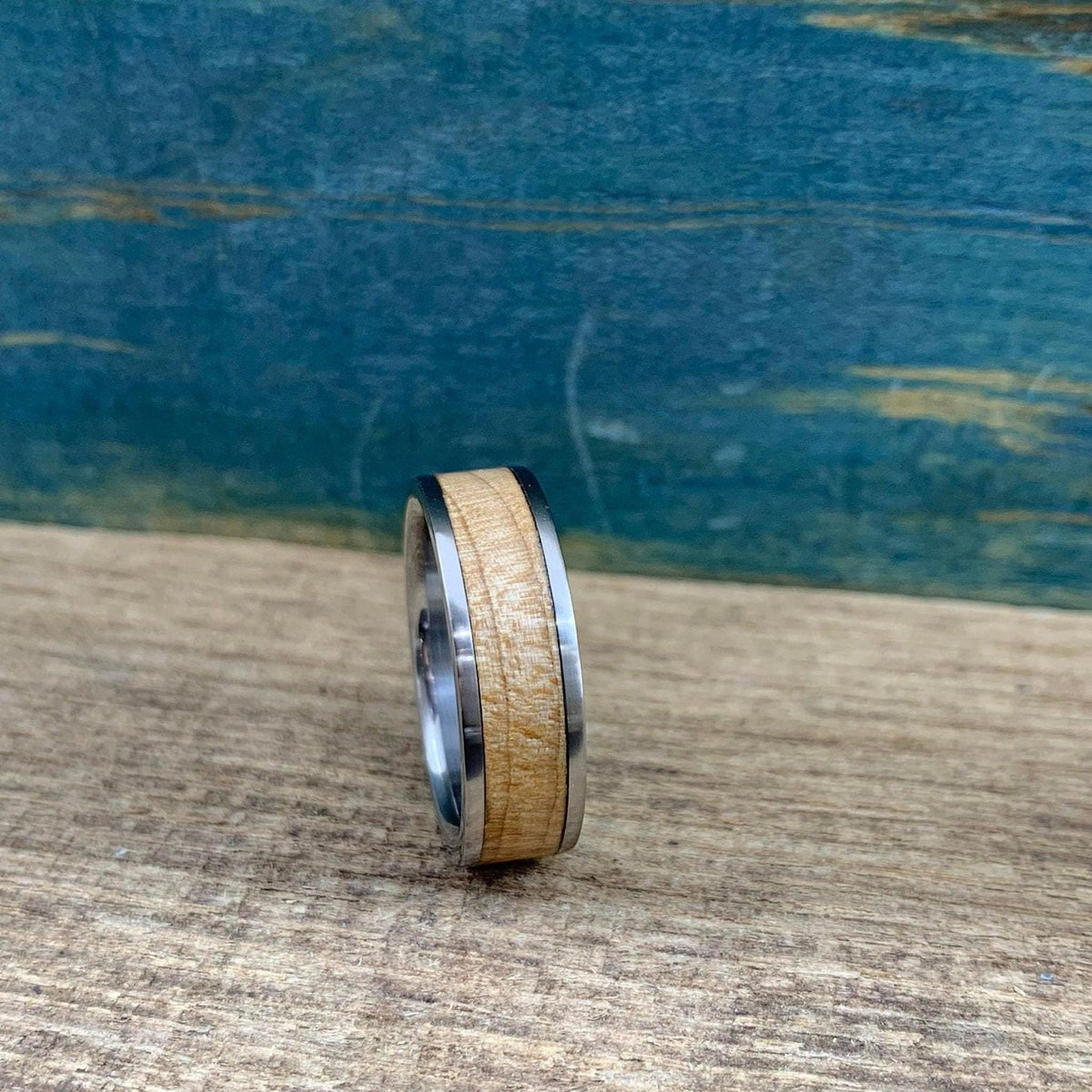 A metal ring with a wide wood inlay stands on a wooden plank against a blue background.