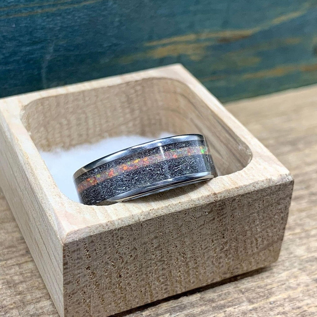 A titanium ring featuring an inlaid band of textured meteorite and a thin center stripe of shimmering champagne opal, resting inside a small open wooden box.