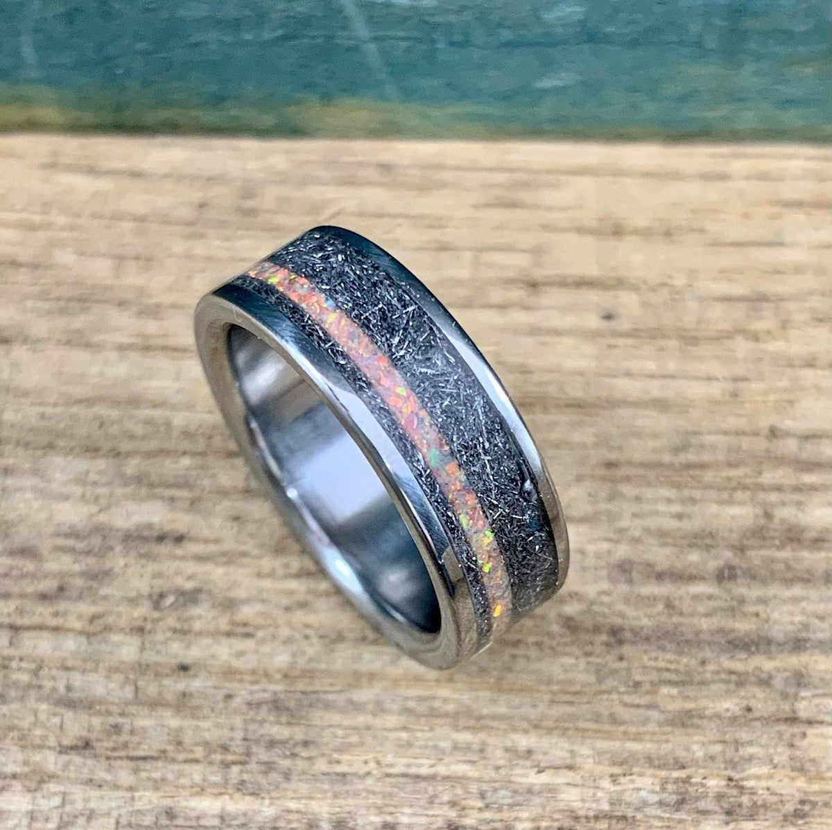 Angled view of a titanium band with a textured dark meteorite inlay bisected by a sparkling peach opal inlay, displayed on a rustic wooden surface.