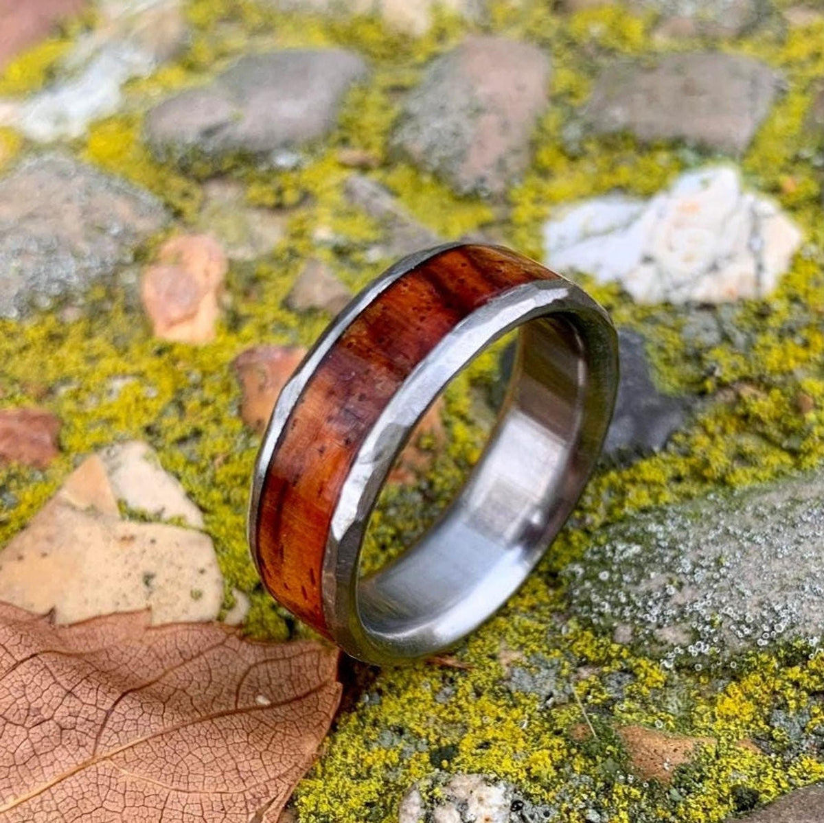 The ring lying diagonally across a dry leaf and mossy ground, showing its textured metal edge.