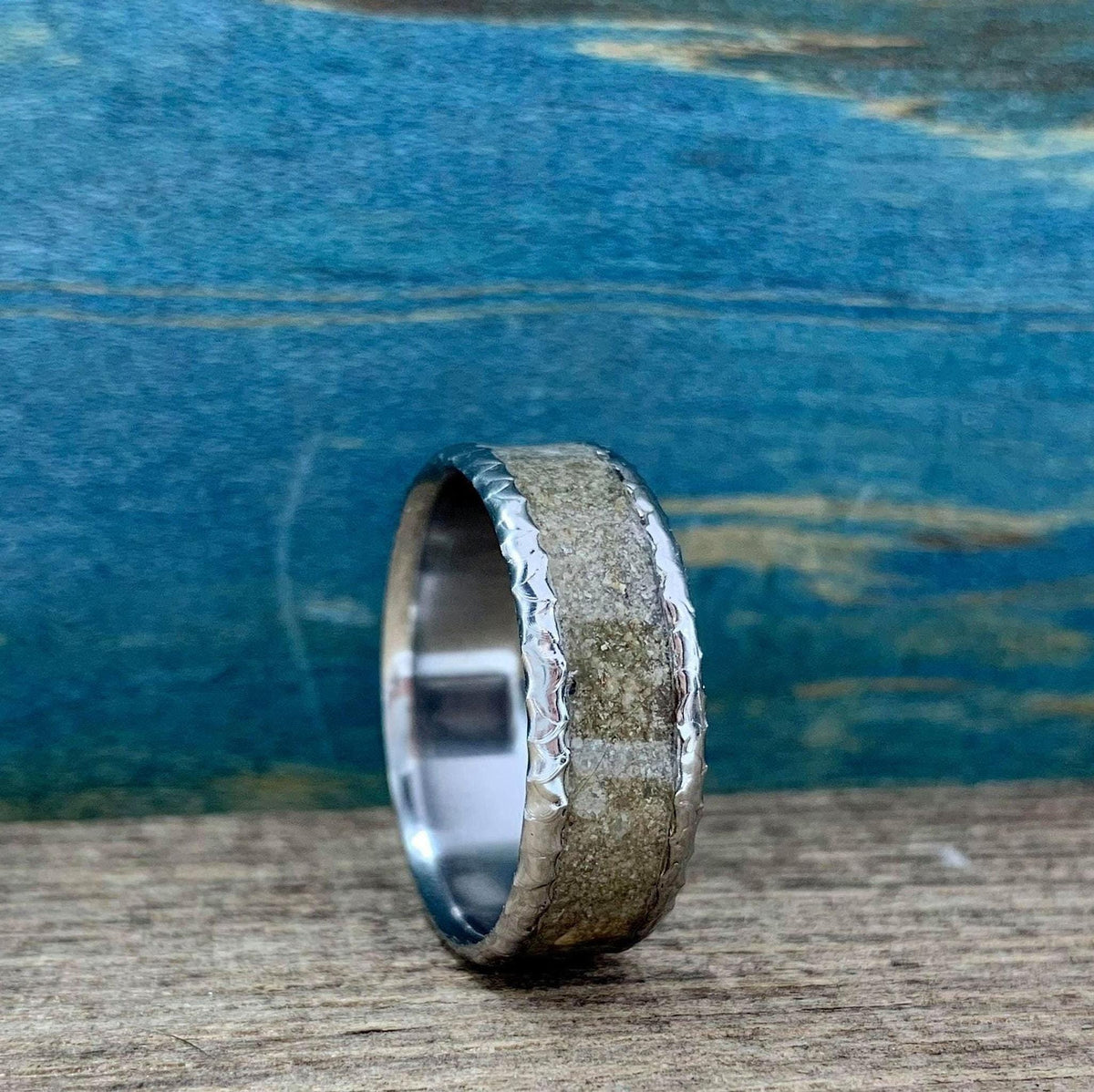 A titanium ring with a textured concrete inlay standing upright on a wooden surface against a blue background.