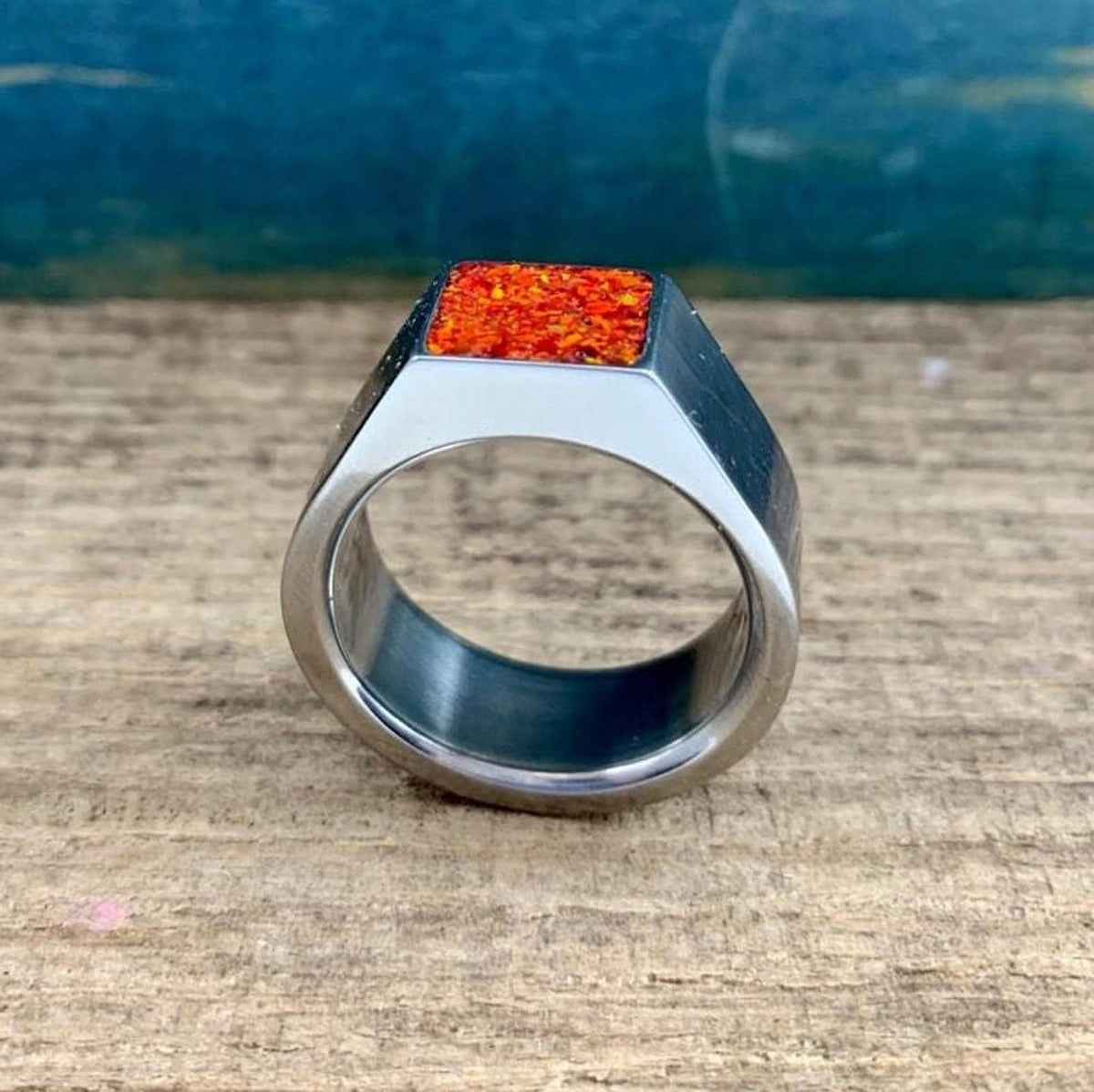 A silver signet ring with a rectangular orange opal inlay rests on a wooden surface against a blue backdrop.