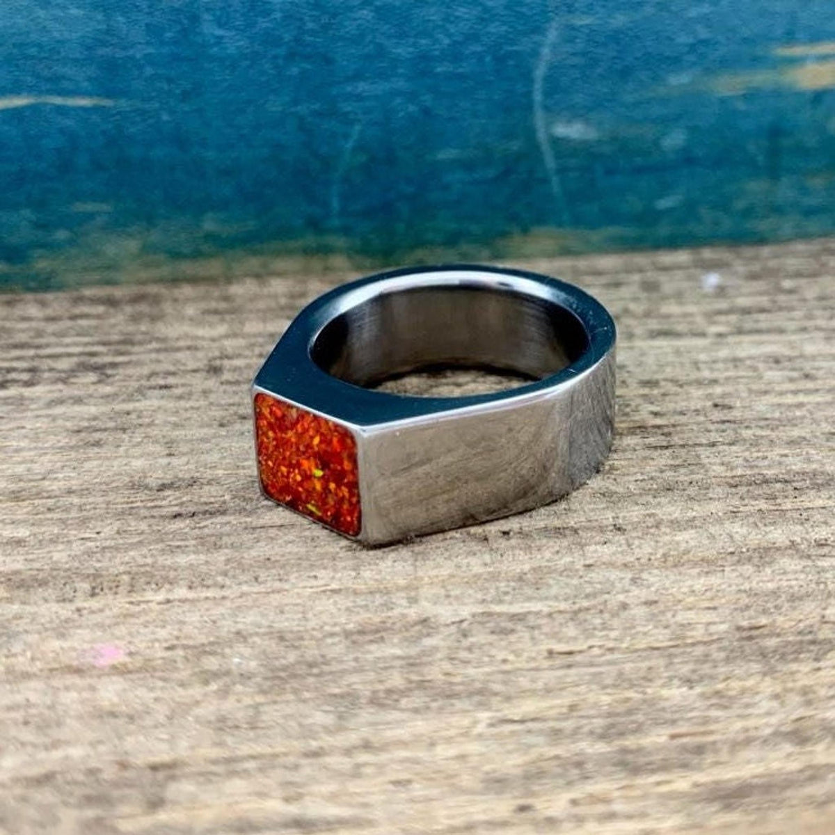 A silver ring with a flat rectangular top containing a fiery orange opal inlay sits on a wooden surface.