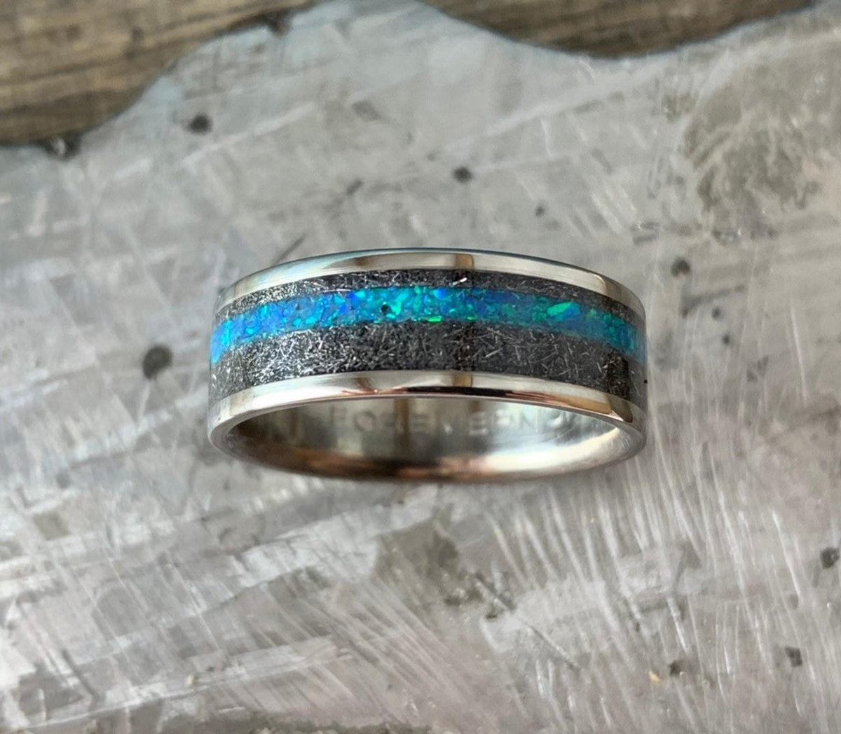The ring lies flat on a light textured surface, showing its bright blue opal stripe bordered by meteorite material.