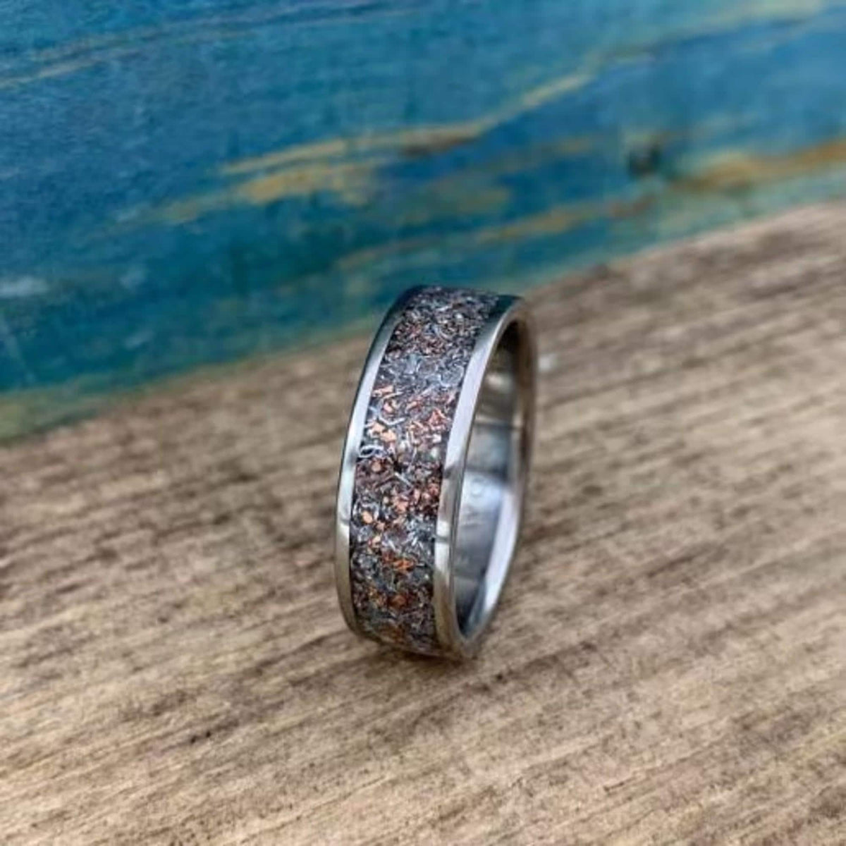 A metal ring with a textured inlay of mixed metallic flakes sitting upright on a wood surface against a blue backdrop.
