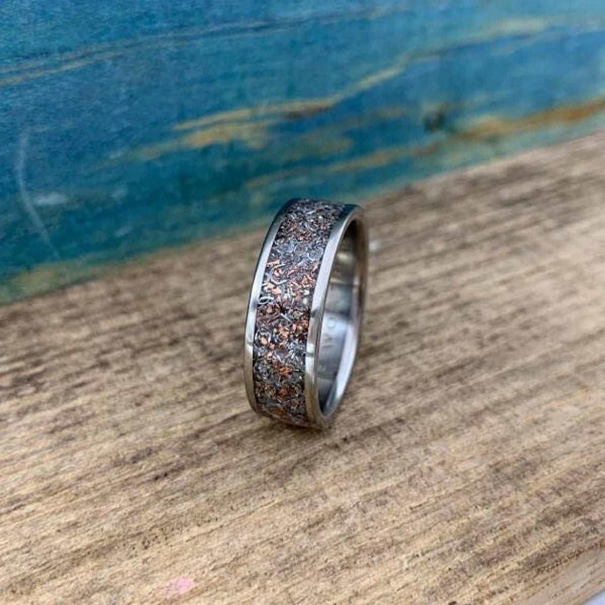 A band ring featuring a wide speckled inlay of copper and silver metal fragments resting on a wooden plank.